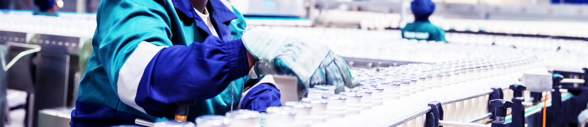 An individual in a blue uniform and head covering is working on a production line in an industrial setting. They are inspecting or handling clear bottles moving along a conveyor belt. Another person in similar attire is visible in the background, also engaged in the production process. The scene is indoors with artificial lighting, showcasing the machinery and the manufacturing workflow.