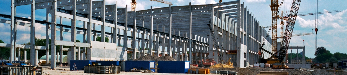 The photo captures a construction site bathed in sunlight. Against a backdrop of a clear blue sky with scattered clouds, a large building is taking shape. The focus is on the steel framework, which stands tall and intricate. Several cranes are actively engaged in lifting materials, emphasizing the industrious nature of the scene. The foreground features dirt and gravel, signifying ongoing groundwork. Overall, it portrays progress, architectural evolution, and the energy of construction.