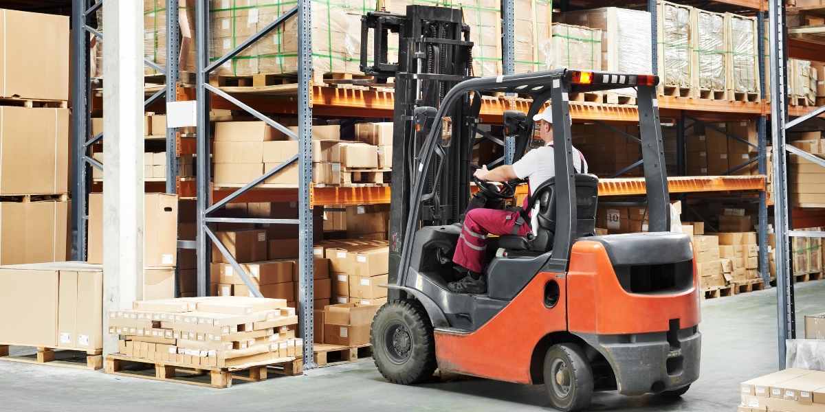 A warehouse forklift operator moving pallets in a large storage facility. The worker is wearing a safety helmet and high-visibility vest, ensuring safe and efficient handling of goods. Search for forklift operator jobs at Aerotek.com